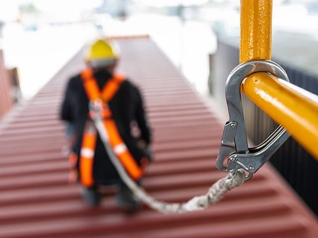 Metal harness clip attached to yellow guardrail is attached by a rope to man in construction hat wearing orange safety harness